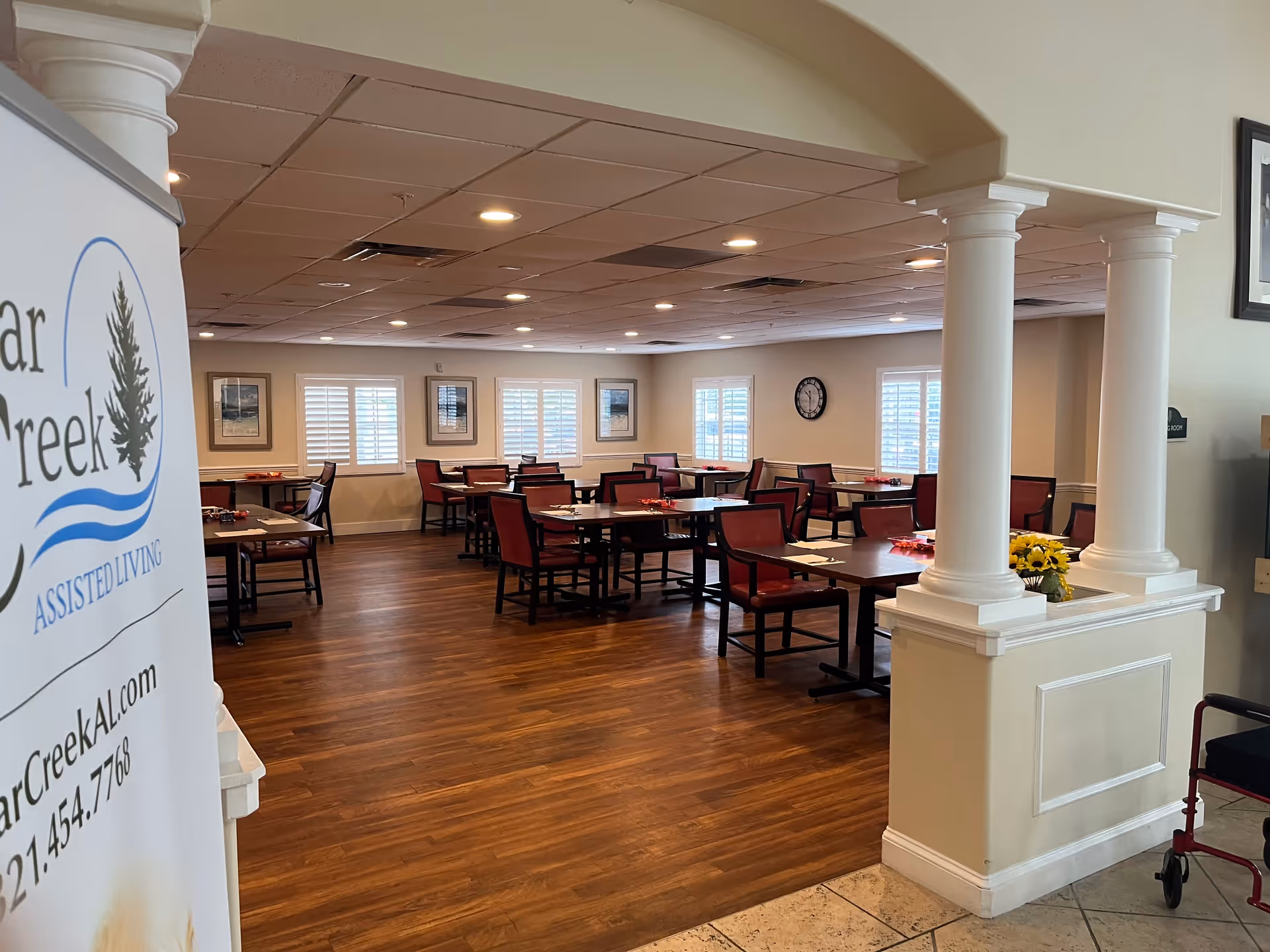 Interior view of a dining room in an assisted living facility with multiple tables and chairs arranged neatly. The room has wooden flooring, white walls, several windows with white shutters, framed pictures on the walls, and a clock. There are decorative columns and a partial view of a banner with Cedar Creek Assisted Living branding on the left side.