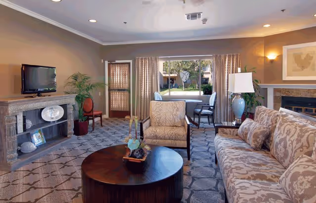 A cozy living room in a senior living facility with a patterned sofa, an armchair, a round wooden coffee table with a small plant, a TV on a stone mantel, a fireplace, and large windows with curtains overlooking a garden area.