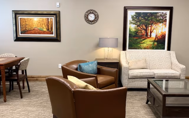 A cozy assisted-living lounge with leather armchairs, a patterned loveseat, glass-topped coffee table, side table with lamp, wall clock, and framed landscape paintings.