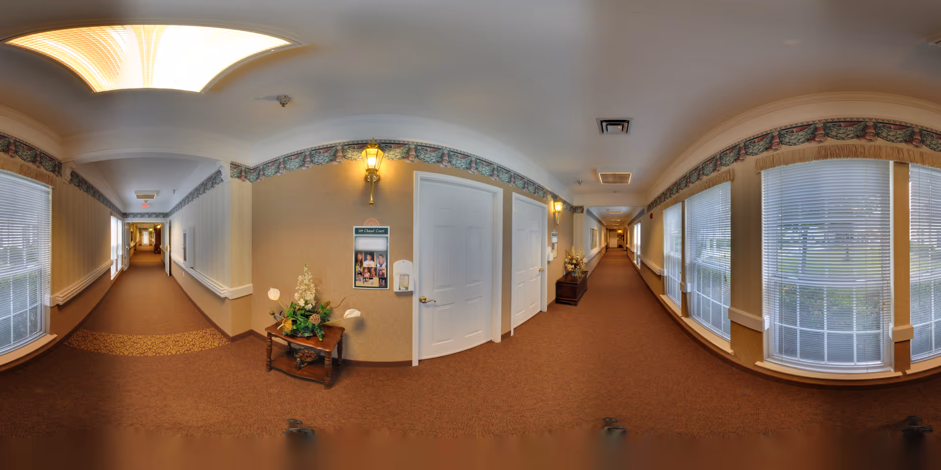 Carpeted corridor lined with large windows, doors, wall sconces and decorative tables in a senior living facility.