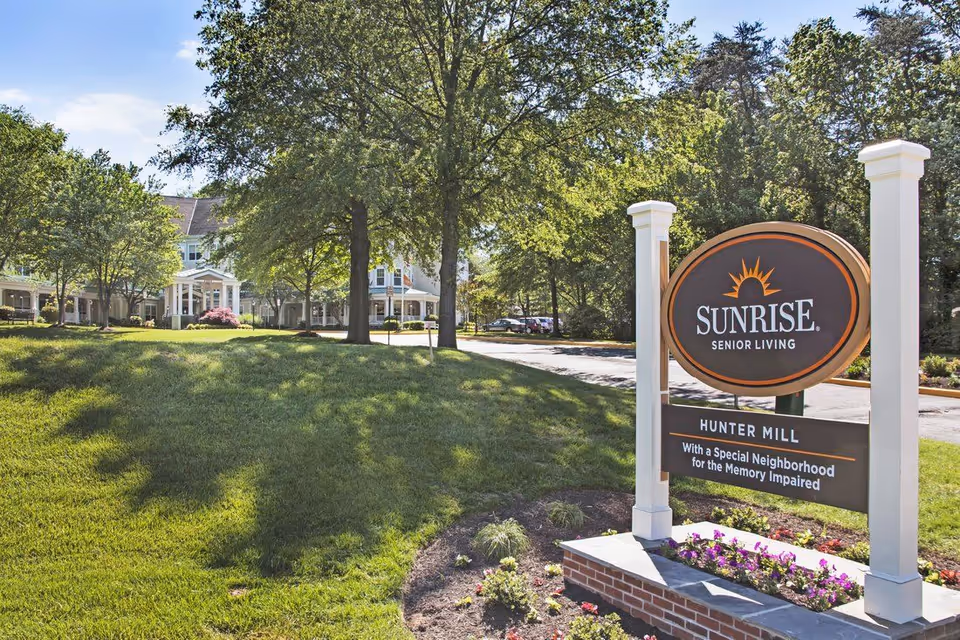 Outdoor view of Sunrise Senior Living at Hunter Mill, featuring a large sign with the facility name and a special neighborhood for the memory impaired. The background shows a well-maintained lawn, trees, and part of the building under a clear sky.