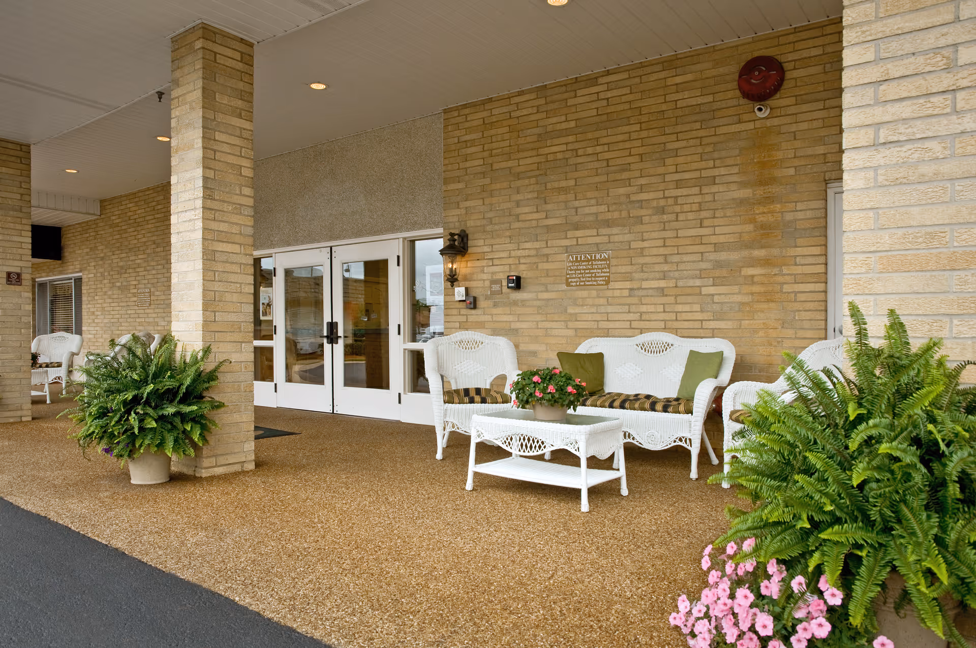Covered brick entrance with white wicker seating, potted plants and double glass doors.