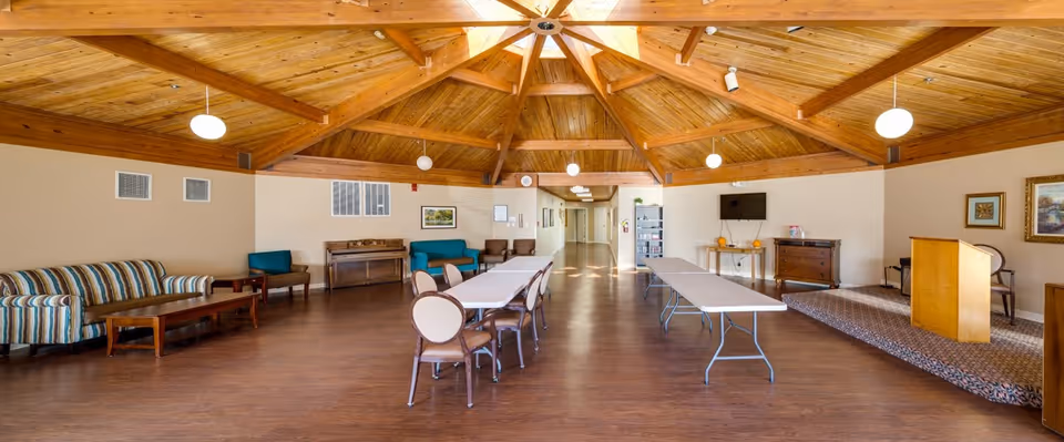 A spacious common area with a high wooden ceiling featuring exposed beams and a skylight. The room has wooden flooring and is furnished with a striped sofa, several chairs, two long folding tables, a podium on a small carpeted platform, a TV mounted on the wall, and various pieces of furniture including a piano and cabinets. The walls are decorated with framed pictures.