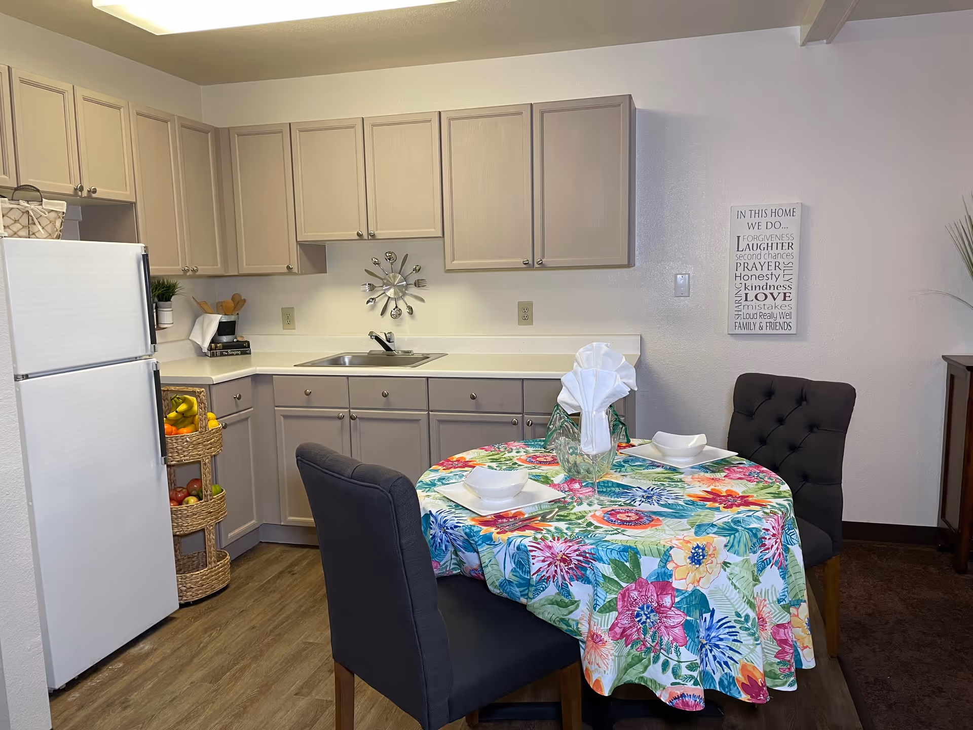 A small kitchen area with beige cabinets, a white refrigerator, and a sink. There is a round dining table covered with a colorful floral tablecloth, set with two white bowls and plates, and two dark upholstered chairs. A decorative wall clock shaped like utensils is mounted above the sink, and a framed sign with inspirational text is on the wall to the right.