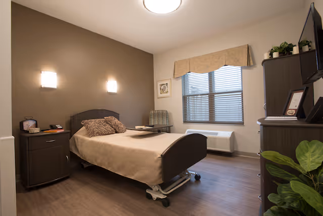 A clean, well-lit senior facility bedroom with a single adjustable bed, nightstand, armchair, dresser with TV, and a window.