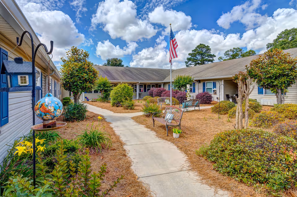 Outdoor courtyard area of a senior living facility with a concrete walkway, benches, various shrubs and plants, an American flag on a flagpole, and a partly cloudy blue sky.