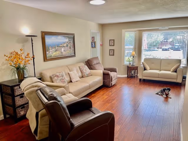 A bright and cozy living room with wooden flooring, featuring a beige sofa with patterned cushions, a brown recliner chair, a cream loveseat near a large window with blinds, a small side table with a lamp and flowers, a tall floor lamp, a framed painting on the wall, and a small dog lying on the floor.