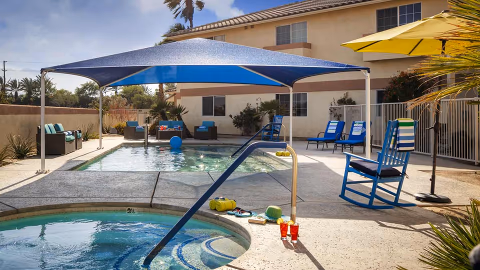 Outdoor pool area at a senior living facility with a shaded swimming pool, lounge chairs, a rocking chair, and a hot tub in the foreground. The area is surrounded by a building and some plants under a clear sky.