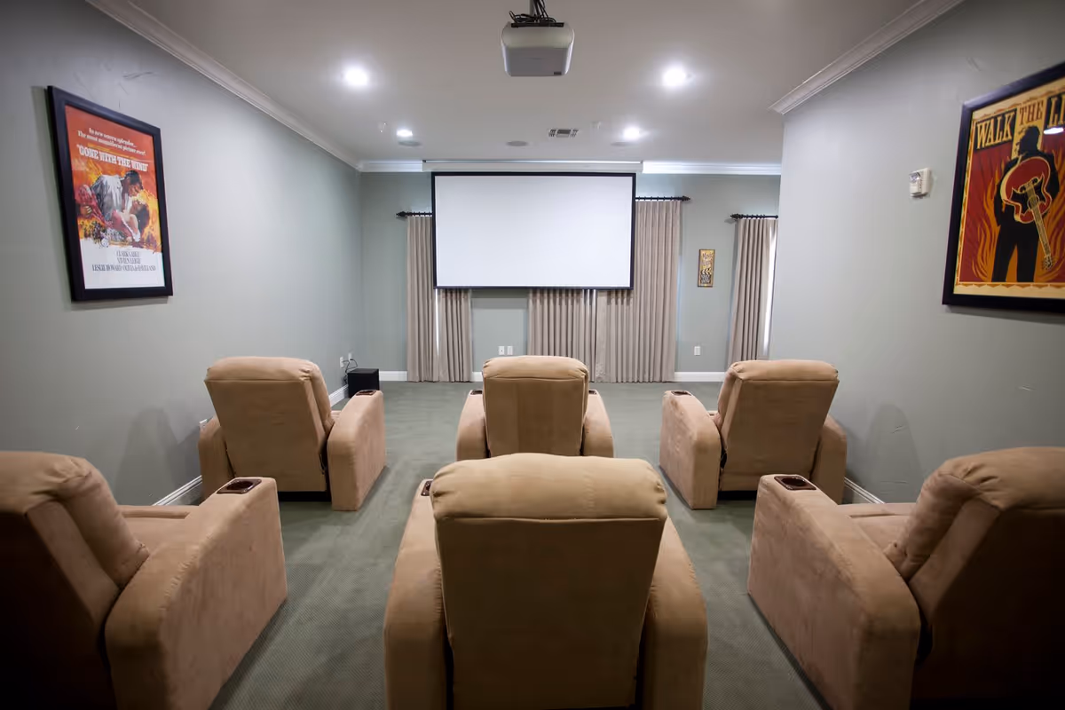 A small home theater room with six beige recliner chairs arranged in two rows facing a large white projection screen. The walls are painted light gray and decorated with framed movie posters. The room has green carpet flooring and ceiling lights.