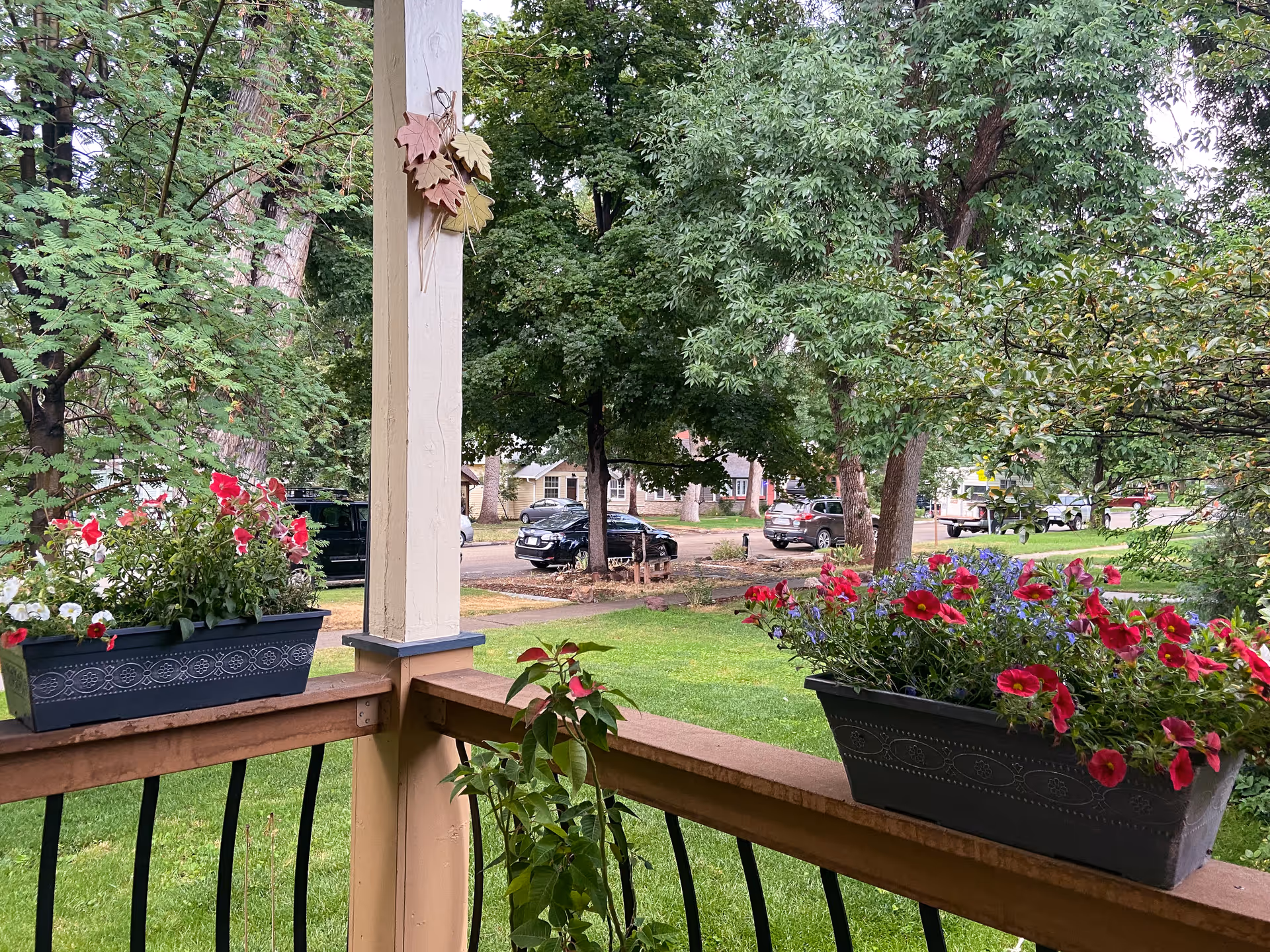 View from a porch with flower boxes containing red, white, and purple flowers. The porch has wooden railings and a white post decorated with three wooden leaves. Beyond the porch is a green lawn, several trees, parked cars, and houses in the background.
