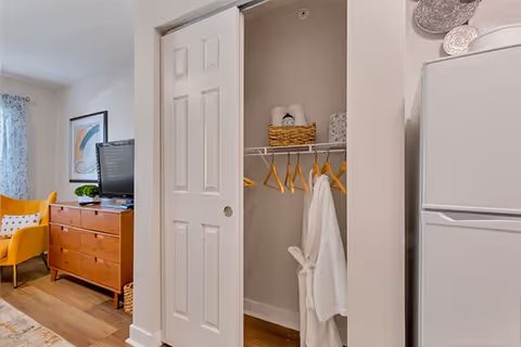 Interior view of a living space in an assisted living facility showing a closet with a sliding door partially open, containing wooden hangers, a white robe, and a basket with towels. To the left, there is a wooden dresser with a TV on top and a yellow armchair beside it. A window with patterned curtains is visible in the background.