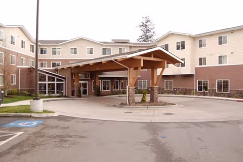 Exterior view of Radcliffe Place senior living facility showing a large covered entrance with wooden beams and stone pillars, surrounded by a multi-story building with beige and brown siding and multiple windows. There is a circular driveway and a handicapped parking space visible in the foreground.