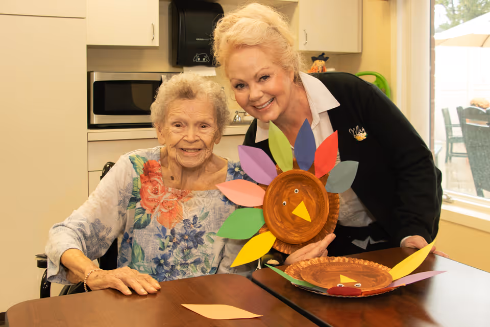 Two smiling women at a table in a care facility holding colorful paper-plate turkey crafts.