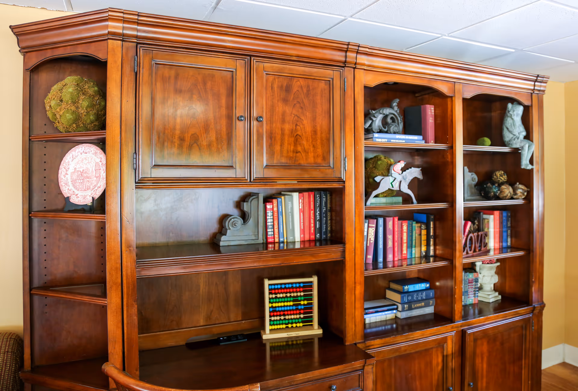 A large wooden bookshelf filled with various decorative items including books, a pink decorative plate, a green moss ball, a wooden horse figurine, an abacus, and other small sculptures, set against a yellow wall.