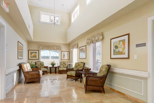A bright and airy sitting area with high ceilings and large windows. The room features six wicker armchairs with floral cushions arranged around two small wooden tables. The walls are painted a soft beige and decorated with framed floral artwork. The floor is tiled with a light brown marble pattern. Window valances match the chair cushions, and there is a chandelier hanging from the ceiling.