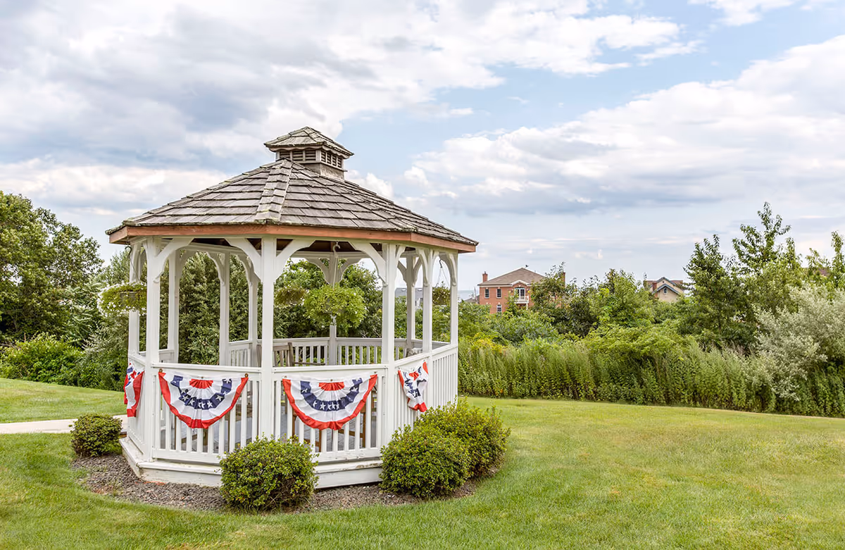 A white wooden gazebo decorated with red, white, and blue patriotic bunting, situated on a well-maintained grassy lawn with bushes around its base. In the background, there are trees and a few houses under a partly cloudy sky.