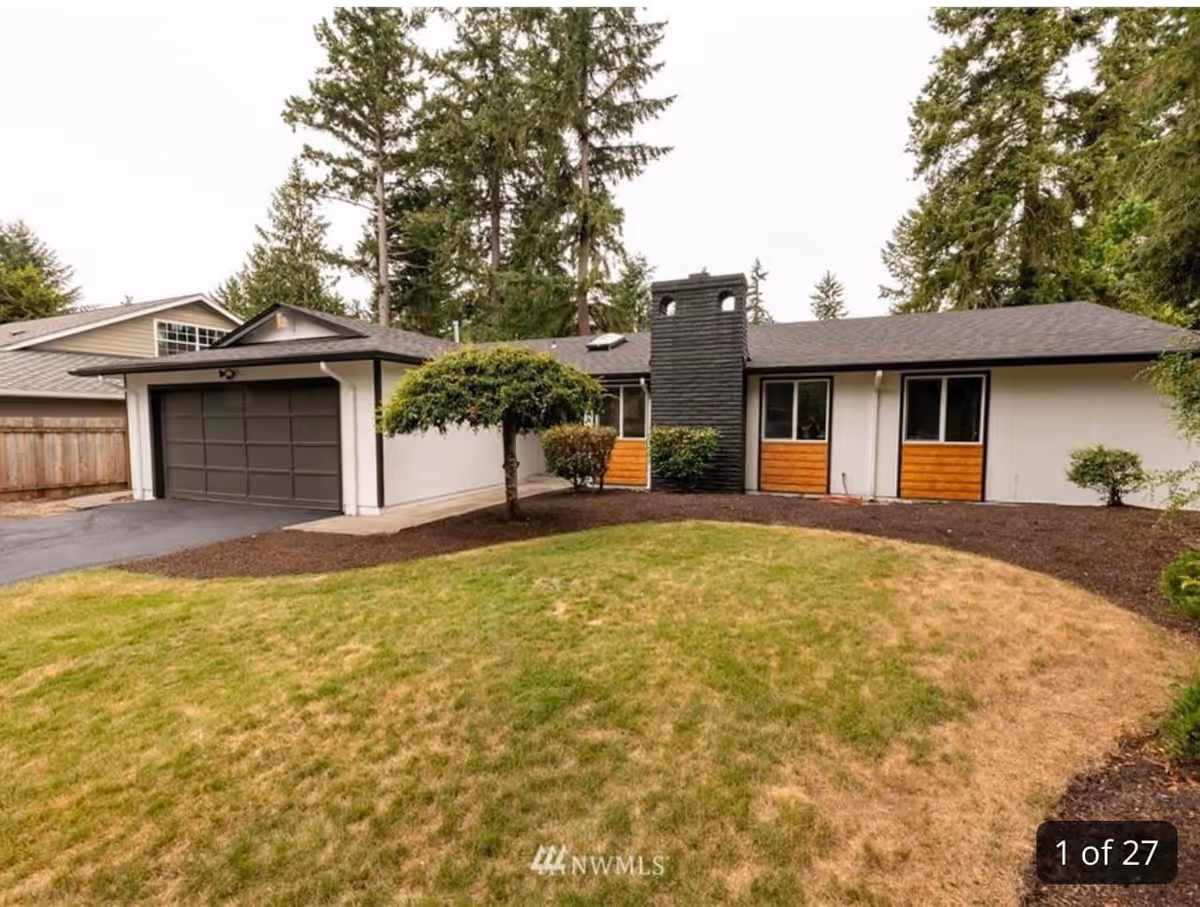 Front exterior view of a single-story house with a dark gray garage door, white walls, and a black chimney. The house is surrounded by a lawn with some patches of dry grass, small bushes, and tall trees in the background.