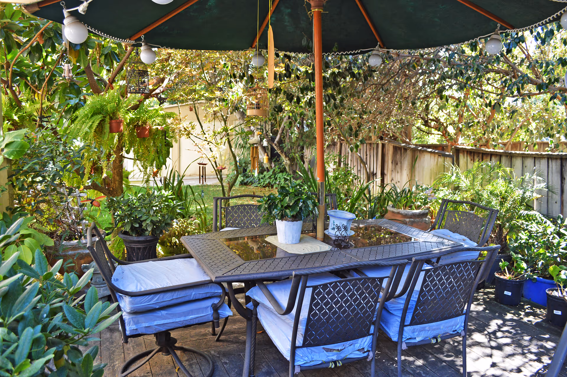Outdoor patio area with a metal table and six cushioned chairs under a large green umbrella. The space is surrounded by lush greenery, potted plants, hanging ferns, and trees, creating a cozy garden atmosphere.