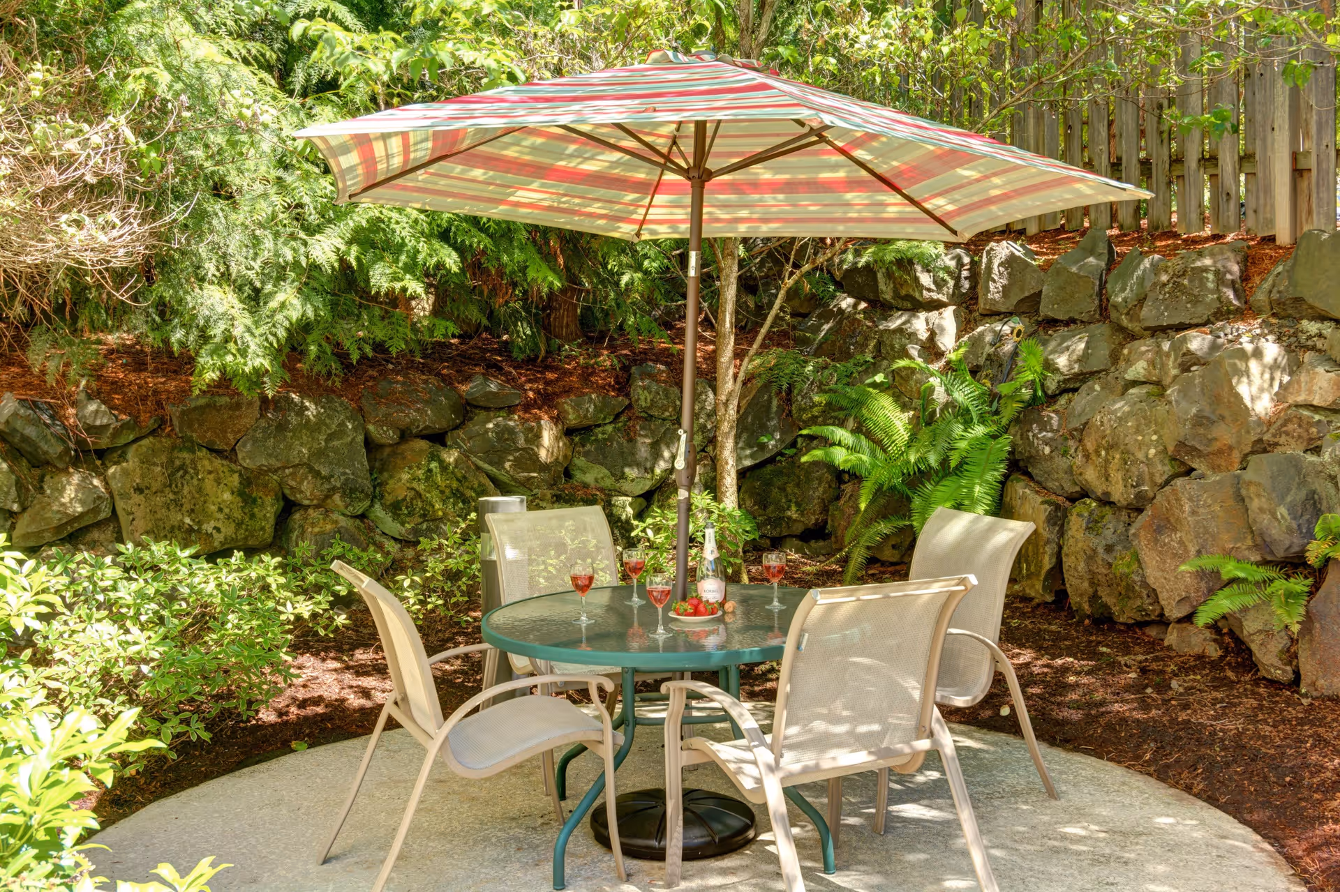 Outdoor patio area with a round glass table surrounded by four beige mesh chairs. A large striped umbrella provides shade over the table. On the table are four glasses filled with a red beverage, a bottle of wine or champagne, and a small plate with strawberries. The patio is bordered by a rock wall and lush green plants and trees.