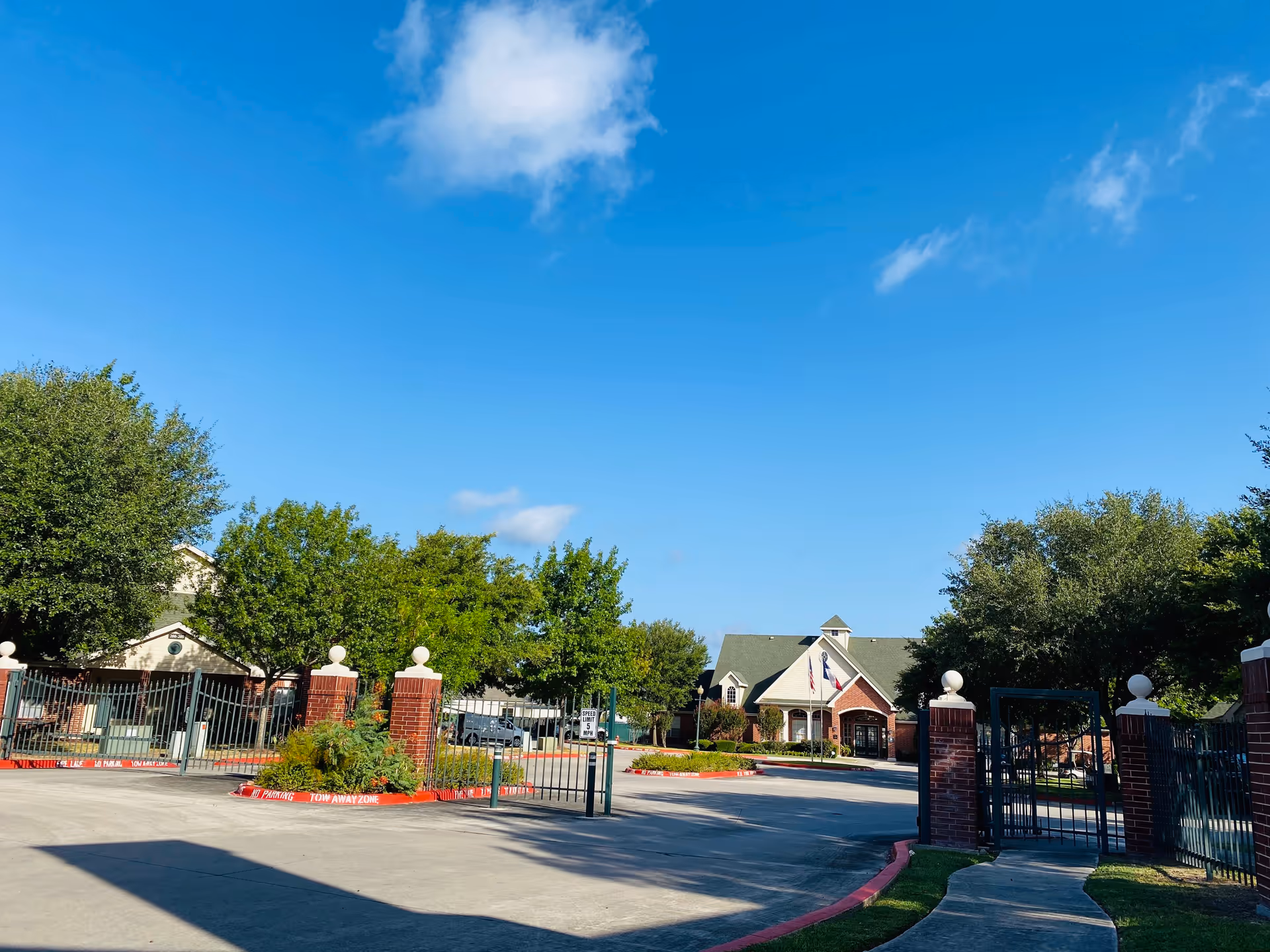 Entrance to The Wellington facility showing a gated driveway with brick pillars and a building with a green roof and flags in the background under a clear blue sky.