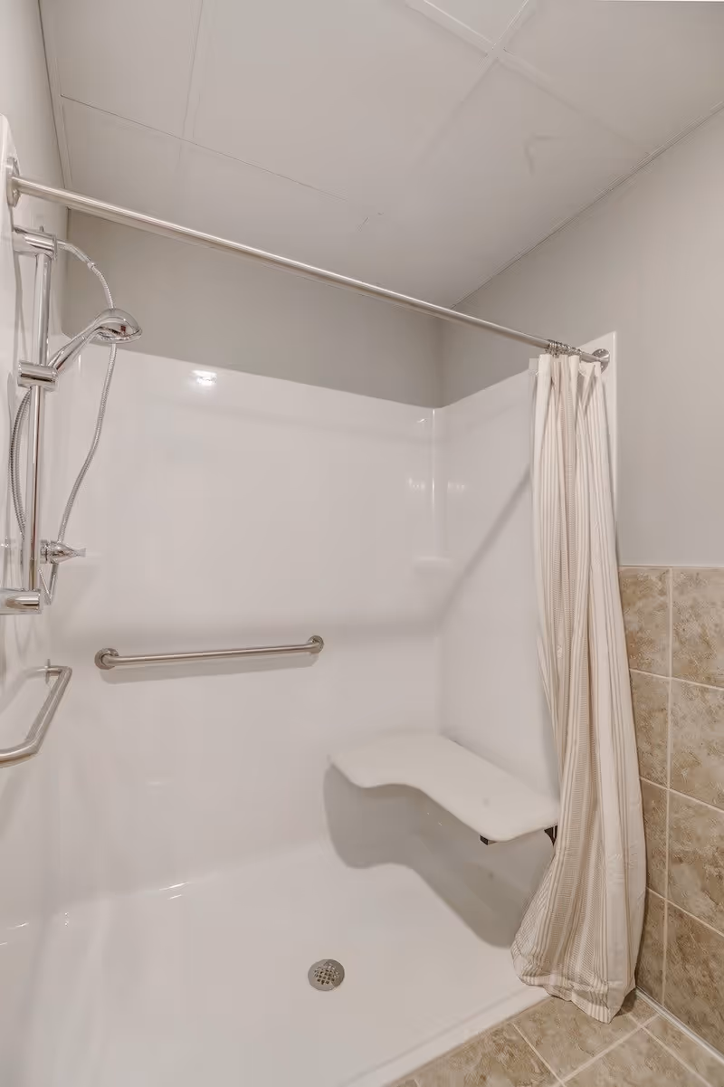 Accessible shower stall with grab bars, a fold-down seat, handheld showerhead, and a shower curtain.