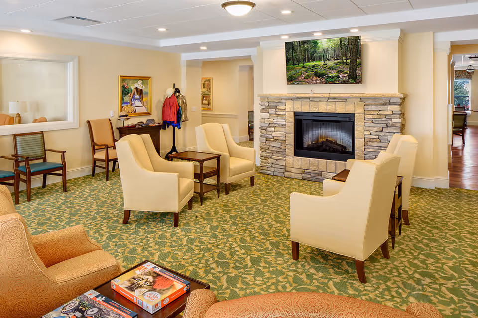 A cozy living room area in a retirement community featuring a stone fireplace with a flat-screen TV mounted above it displaying a forest scene. The room has beige walls, a green patterned carpet, and several beige upholstered armchairs arranged around small wooden tables. There is a coat rack with jackets and hats, a painting on the wall, and a window looking into another room.