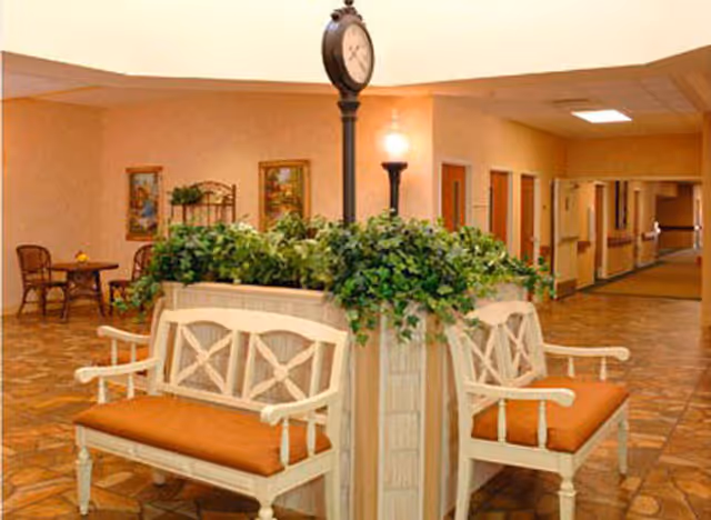 Indoor senior living facility seating area with benches arranged around a central planter topped by a tall clock in a carpeted hallway.