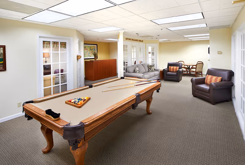 Well-lit community lounge with a pool table in the foreground and seating and tables in the background.