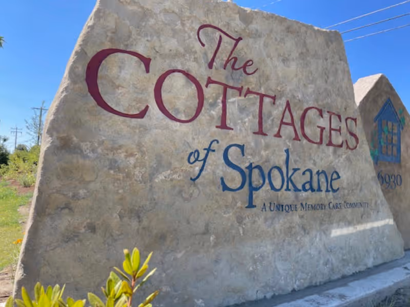 Large stone sign outdoors with the text 'The Cottages of Spokane A Unique Memory Care Community' and a small house logo with the number 6930, set against a clear blue sky.
