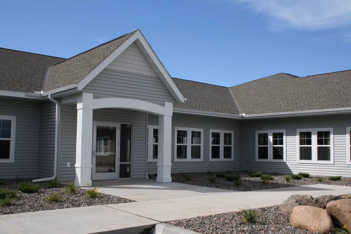 Single-story light-gray senior living building exterior showing the entrance with a gabled portico and walkway.