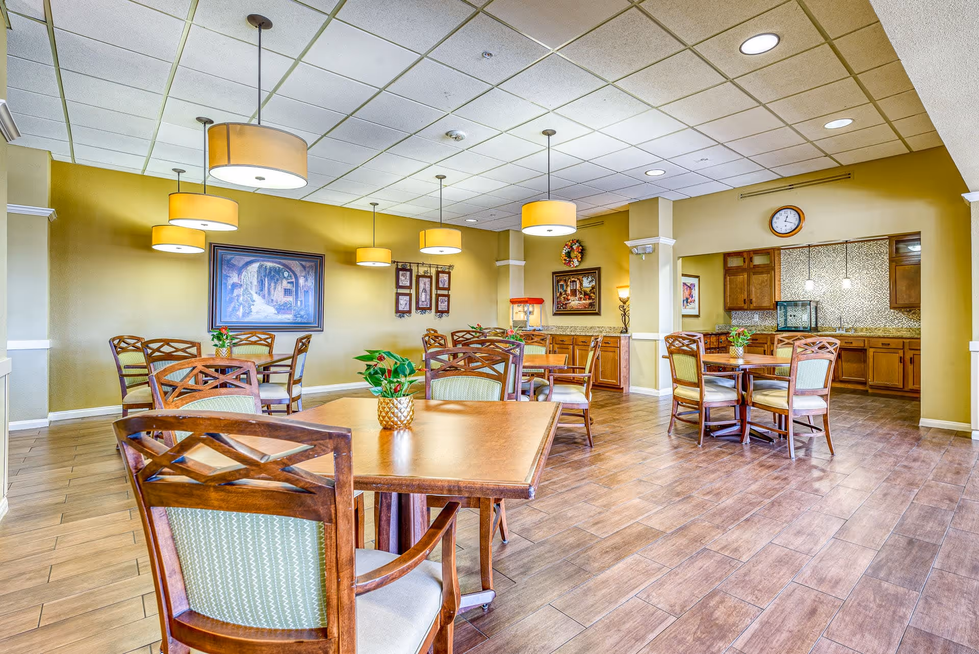 Spacious senior dining room with wooden tables and chairs, pendant lights, and a serving area/kitchen in the back.