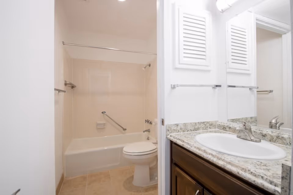 A clean bathroom featuring a bathtub with beige tiles, a white toilet, and a vanity with a granite countertop and a sink. There is a large mirror above the sink and a towel bar on the wall.