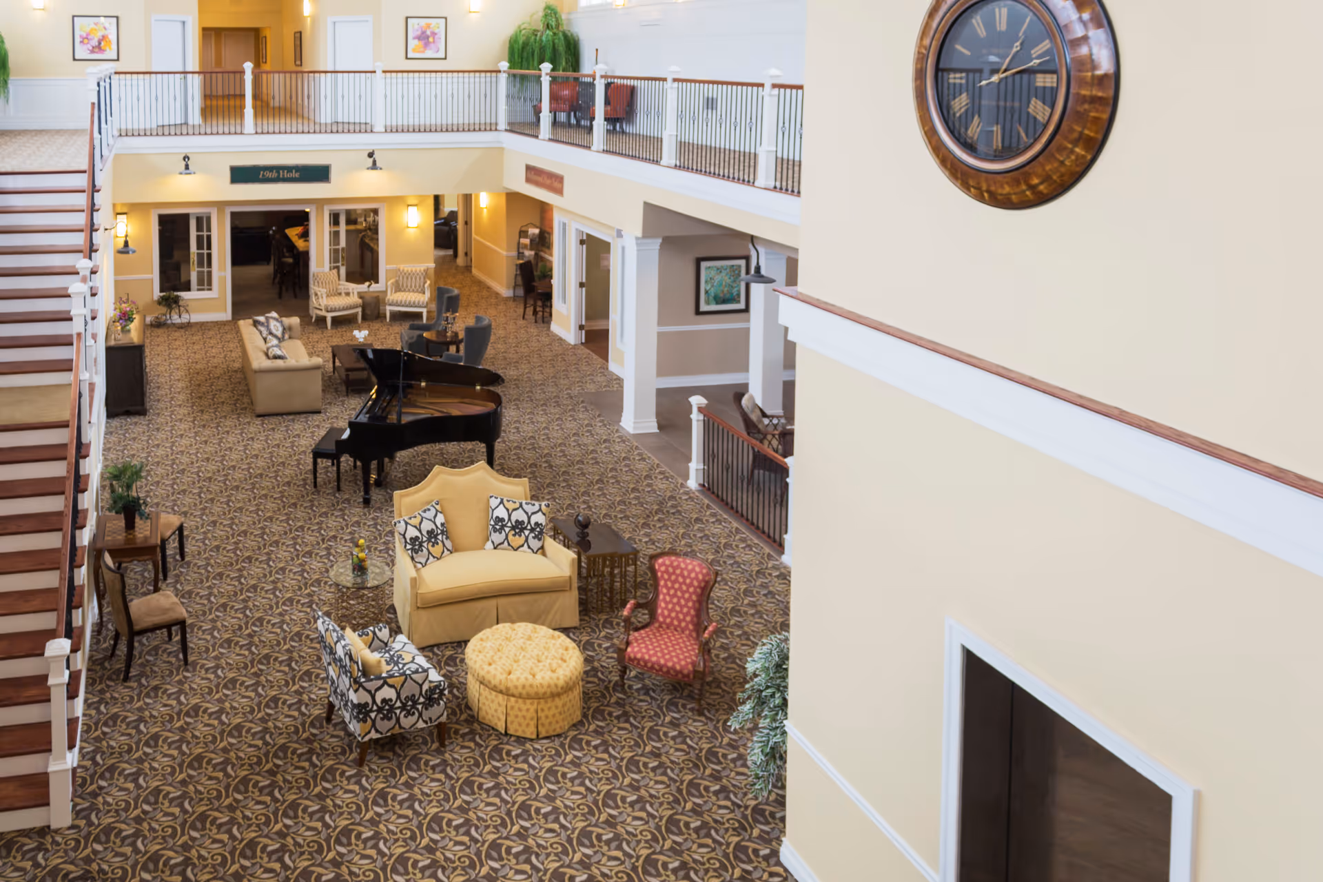 Spacious senior living facility common area with patterned carpet, various seating arrangements including armchairs, sofas, and a yellow loveseat with decorative pillows. A black grand piano is centrally placed. The area has a two-story ceiling with a large wall clock and a staircase on the left side leading to an upper balcony with railings. The walls are painted in light yellow and beige tones, with framed artwork and wall sconces providing lighting.