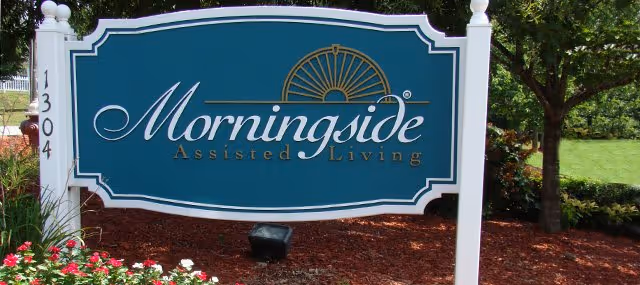 A teal sign reading "Morningside Assisted Living" displayed in landscaped flower beds with trees in the background.