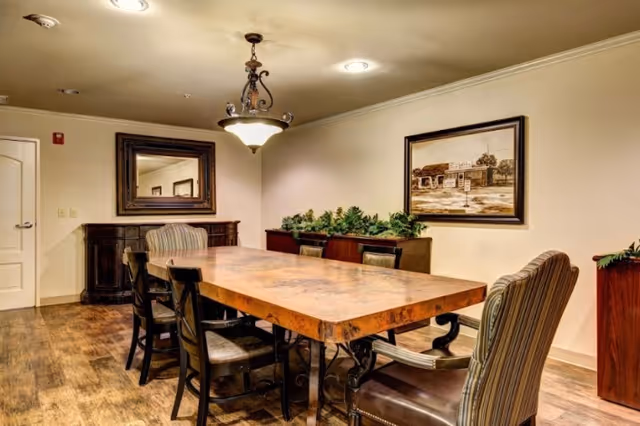 Communal dining room with a long wooden table surrounded by chairs, a chandelier overhead, and artwork and a mirror on the walls.
