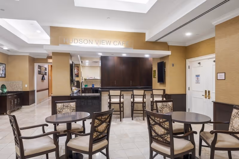Interior view of the Hudson View Cafe at Five Star Premier Residences of Yonkers, featuring round tables with cushioned chairs arranged in the seating area. The cafe counter with high chairs is visible in the background, along with warm beige walls and tiled flooring.