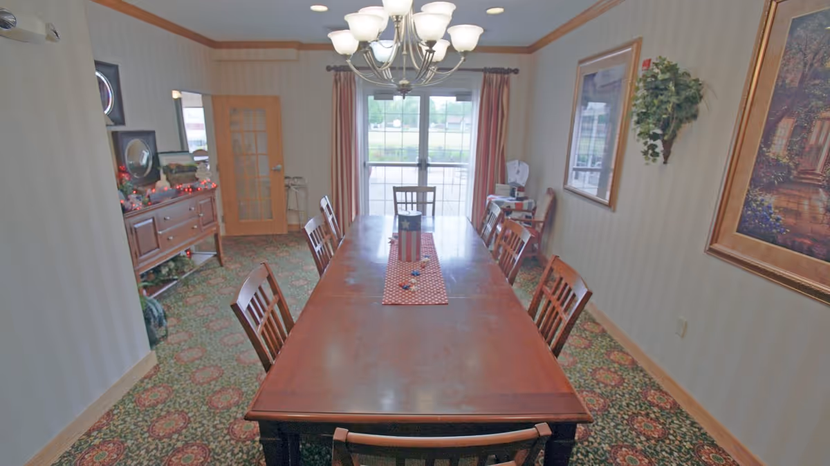 A dining room with a long wooden table surrounded by eight wooden chairs. A red and white table runner with small decorative items is placed on the table. The room has patterned carpet flooring, beige striped wallpaper, and a chandelier hanging above the table. There are framed paintings and a wall-mounted plant on the right wall. At the far end, there are glass doors with red curtains leading outside. On the left side, there is a wooden sideboard with decorations and a wooden door with glass panels.