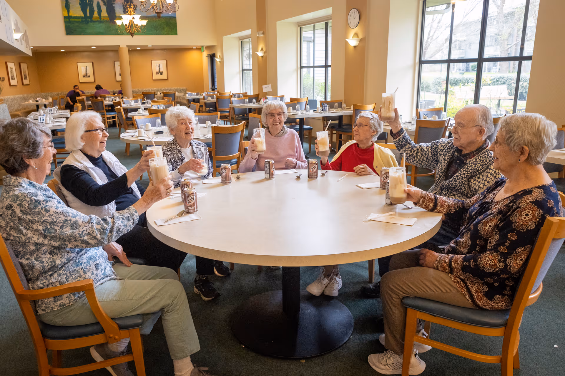 Seven elderly people sitting around a large round table in a dining room, raising glasses filled with a creamy beverage in a toast. The room has large windows letting in natural light, and there are empty tables and chairs in the background.