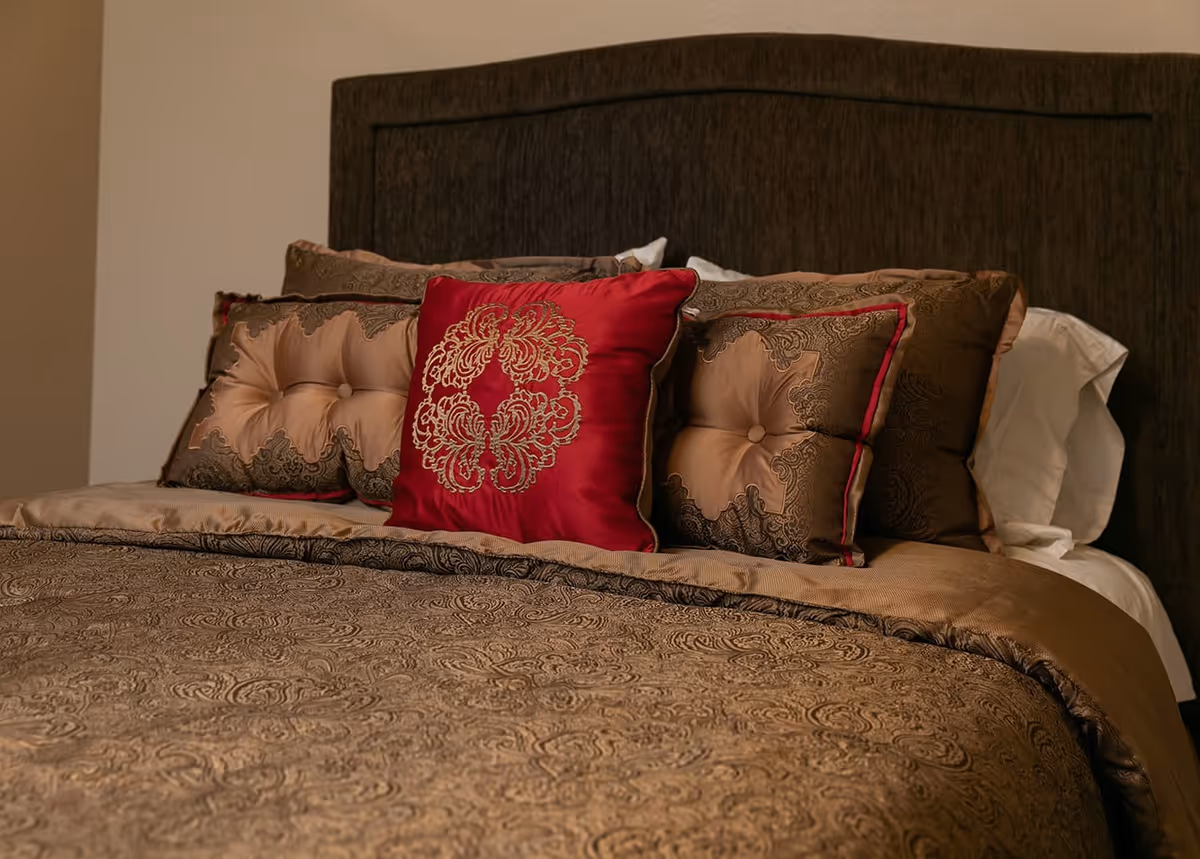 A neatly made bed with an ornate dark wooden headboard, decorated with multiple pillows including a prominent red pillow with gold embroidery, and a brown patterned bedspread.