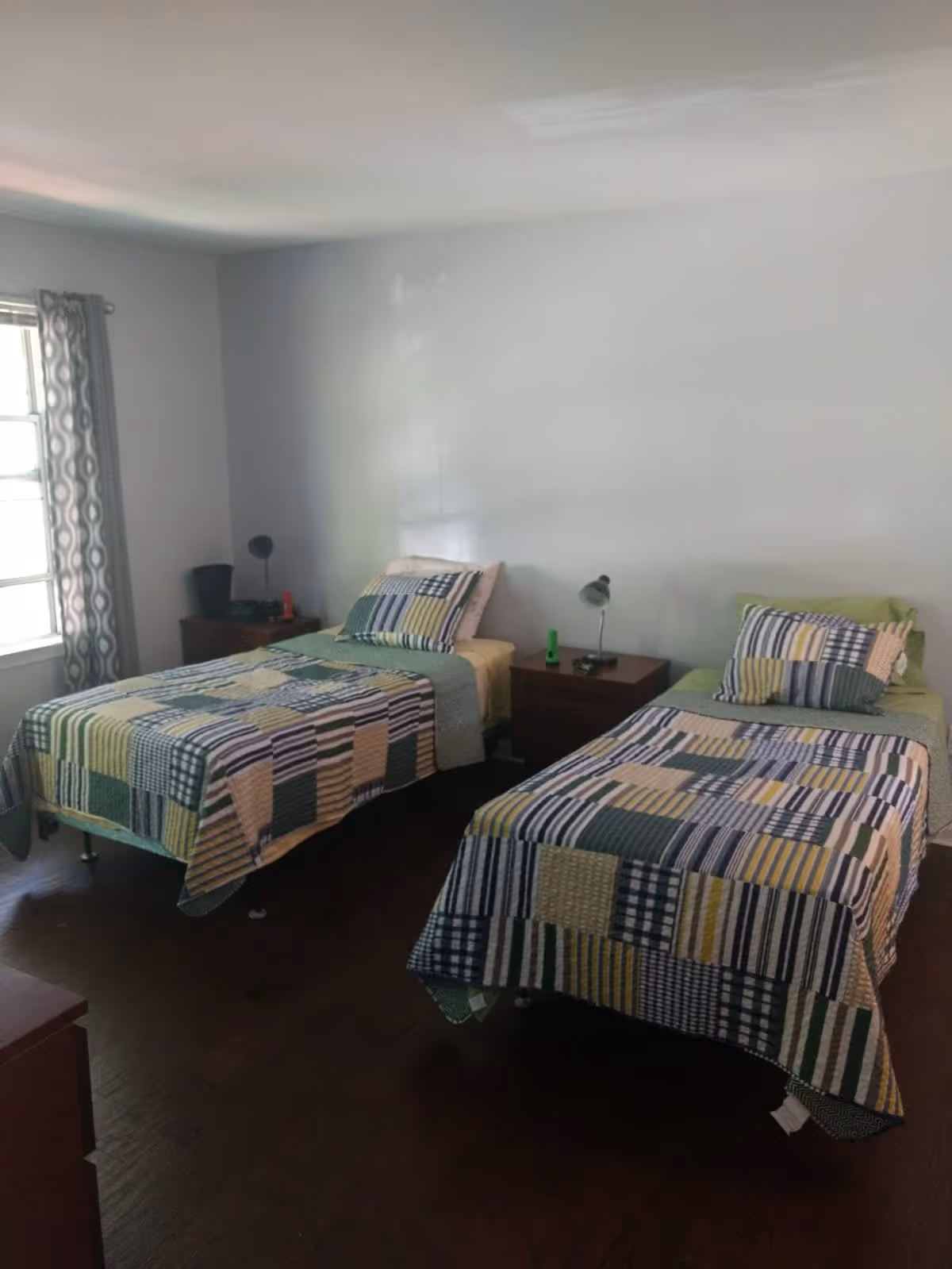 A simple bedroom with two twin beds dressed in patterned quilts, a central nightstand with lamps, and a window with curtains.