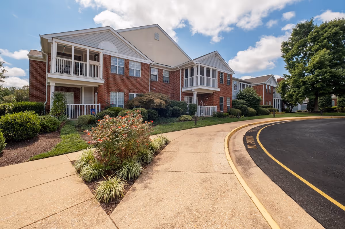 Exterior view of a two-story brick building with white balconies and railings, surrounded by well-maintained landscaping including bushes, flowers, and trees under a partly cloudy sky.
