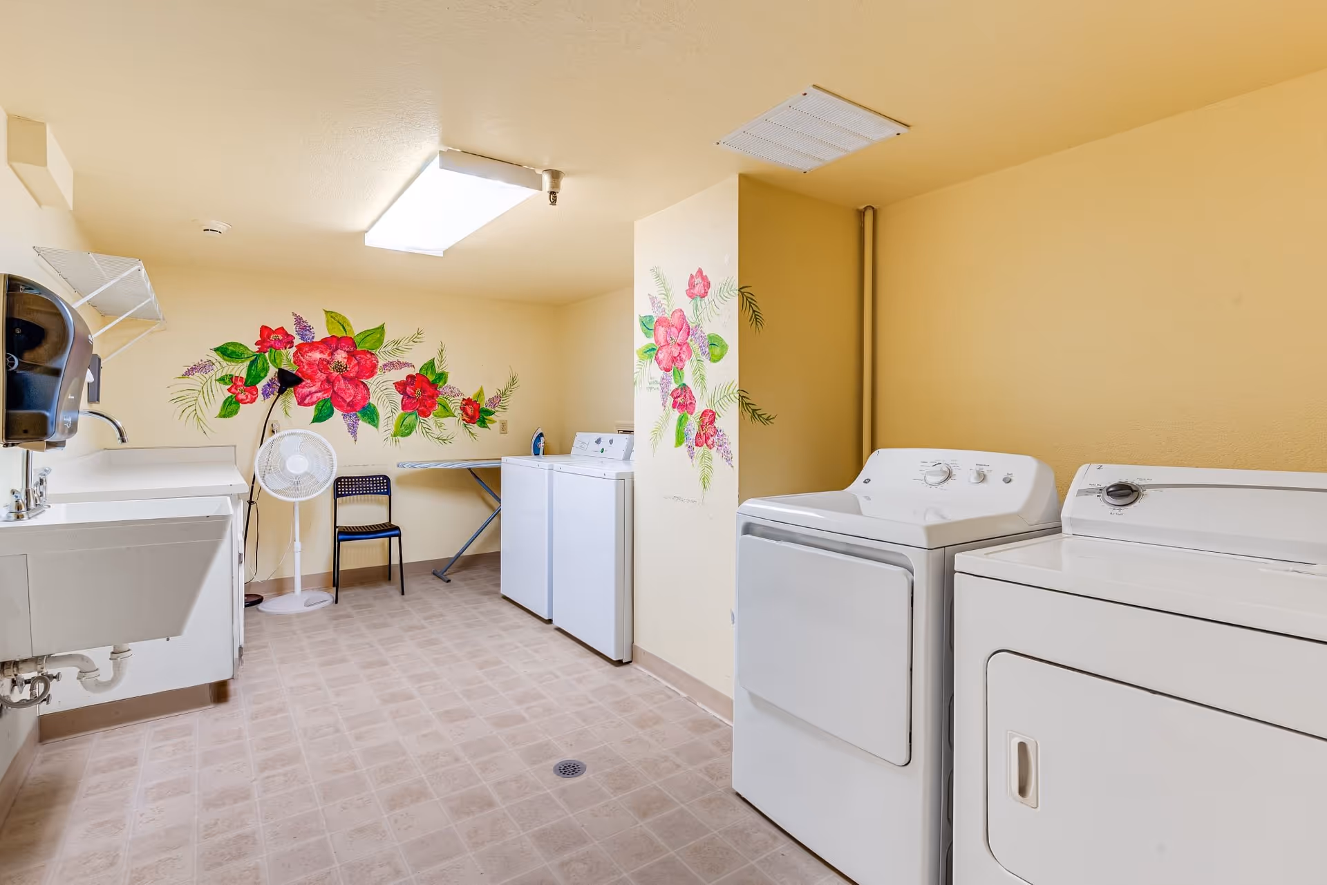 Laundry room with two white washing machines and two white dryers, a utility sink, an ironing board, a chair, a standing fan, and floral wall decorations on yellow walls.
