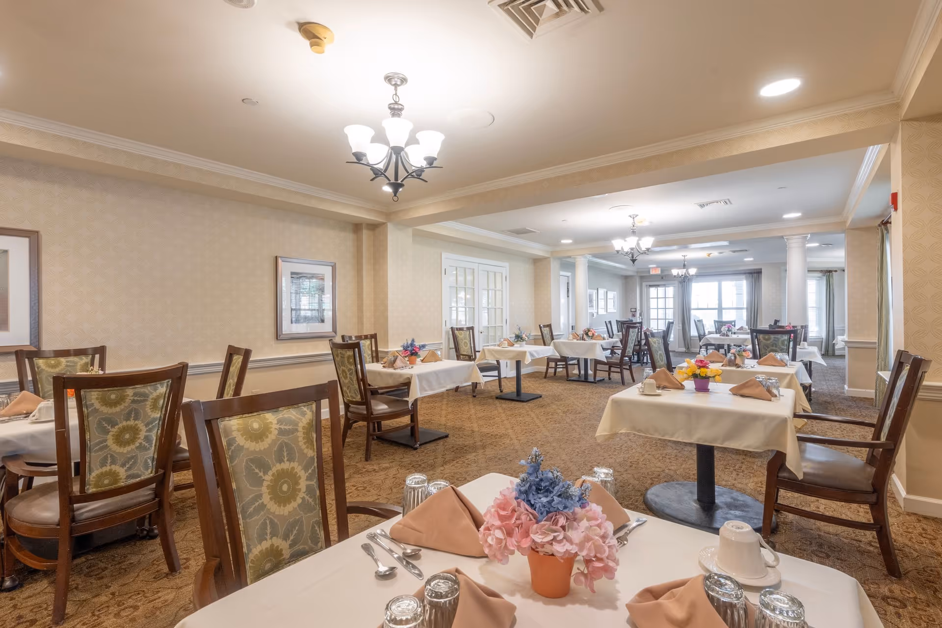 Bright, formal dining room with multiple set tables, chairs, and floral centerpieces.