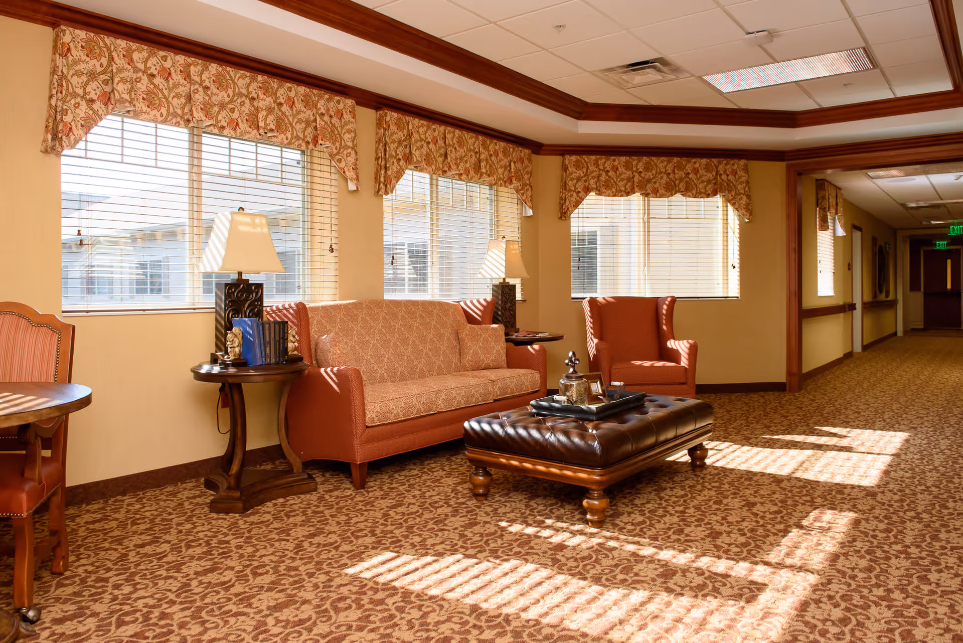 A well-lit sitting area in a senior living facility with a patterned carpet, a sofa, an armchair, a leather ottoman with decorative items on top, two side tables with lamps, and large windows with floral valances letting in sunlight.