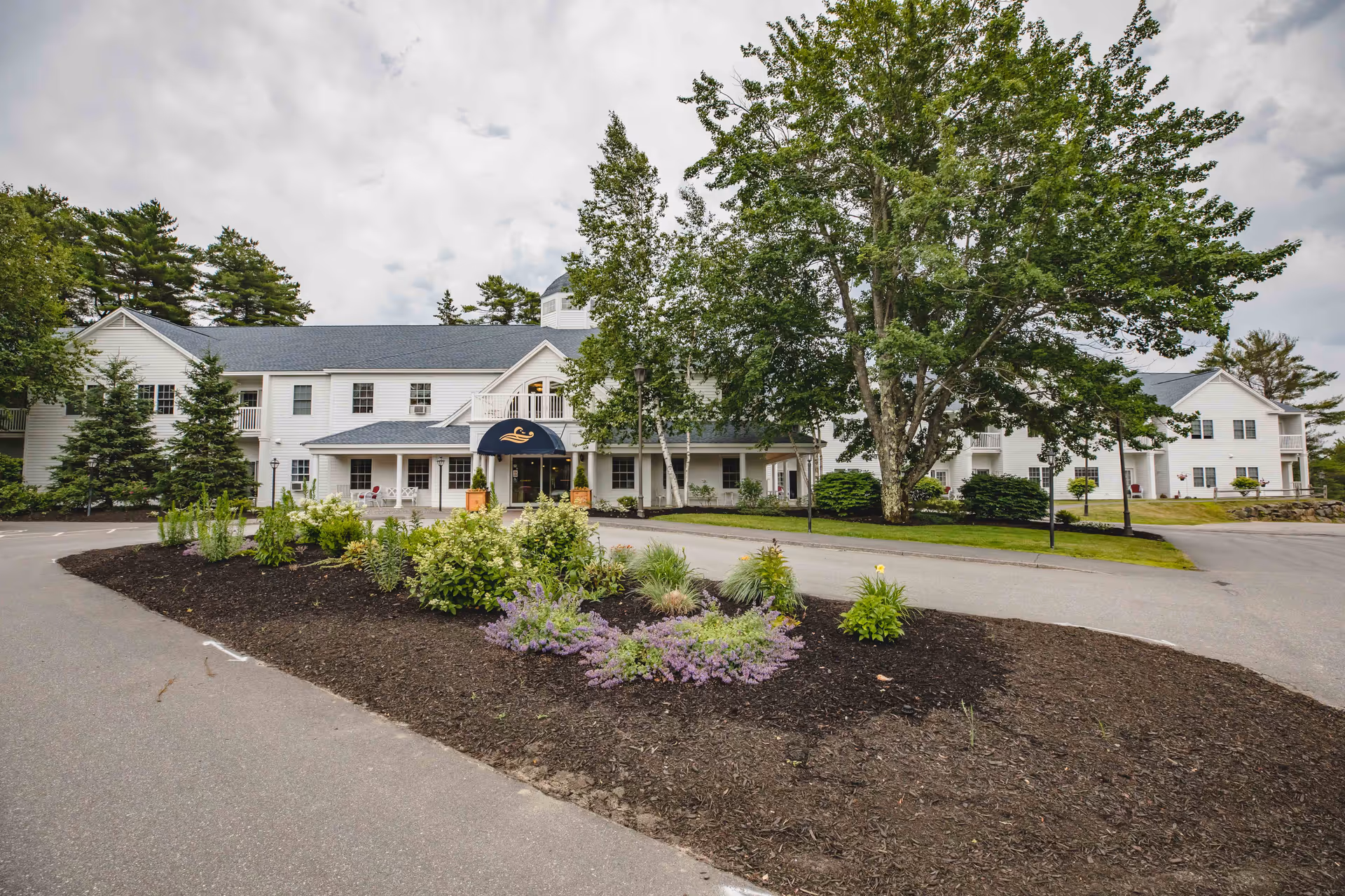 Exterior view of a large white senior living facility building with multiple windows and a dark gray roof. The entrance has a blue awning with a logo, and there is a landscaped area with various green plants and purple flowers in the foreground. Tall trees surround the building under a cloudy sky.
