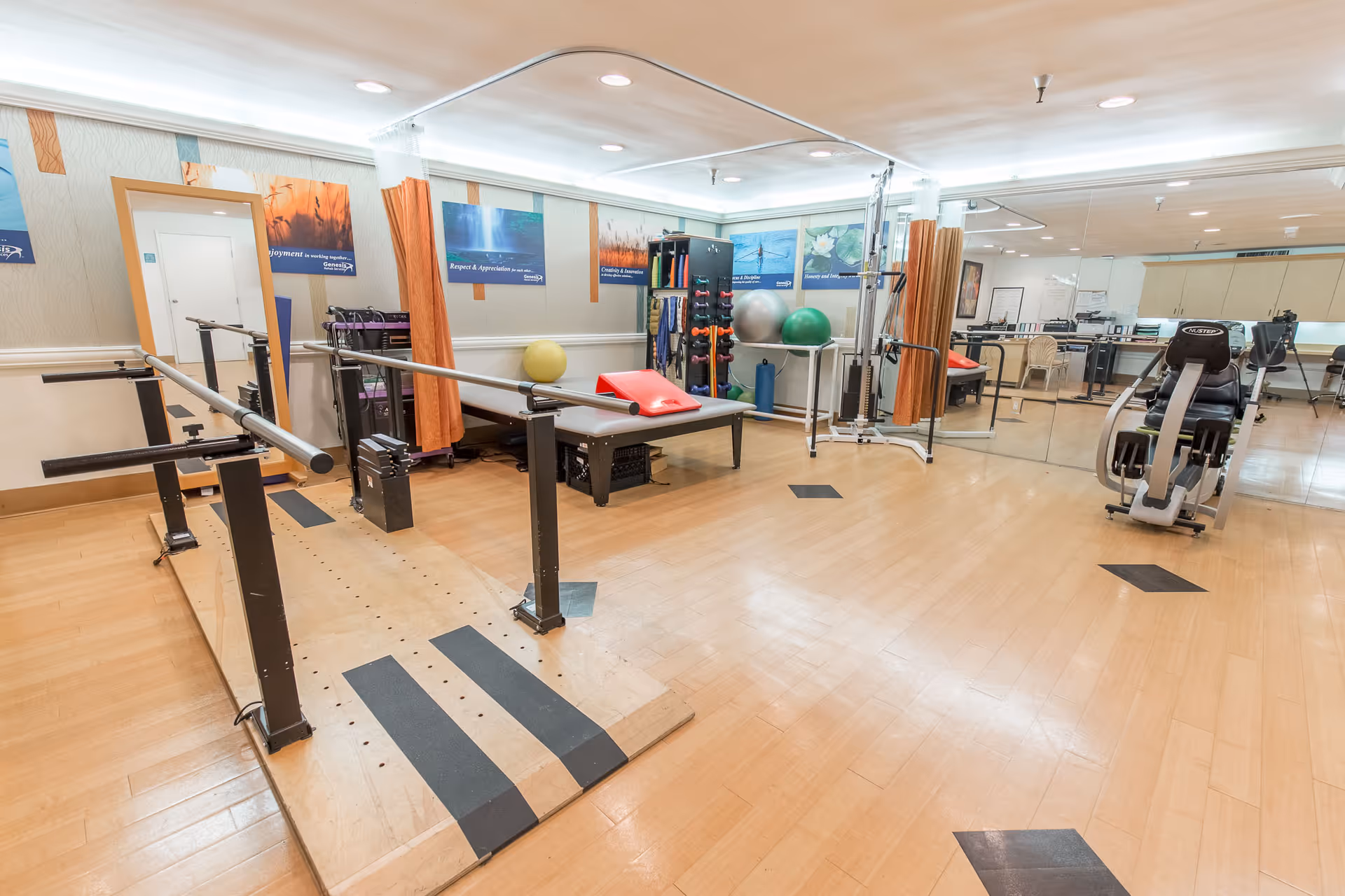 A rehabilitation or physical therapy room with wooden flooring, parallel bars for walking exercises, exercise balls, weights, a therapy table with a red cushion, and various exercise equipment. The room has large mirrors on one wall and motivational posters on the opposite wall.