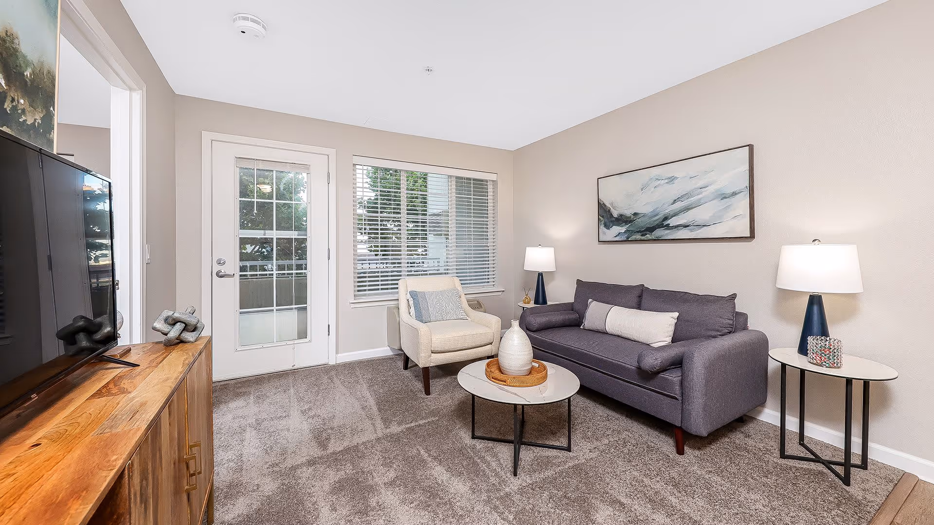 A cozy living room with a gray sofa, a beige armchair, two round coffee tables with decorative items, two table lamps on side tables, a large flat-screen TV on a wooden cabinet, a window with blinds, and a glass door leading outside. The walls are light-colored and there is a large abstract painting above the sofa.