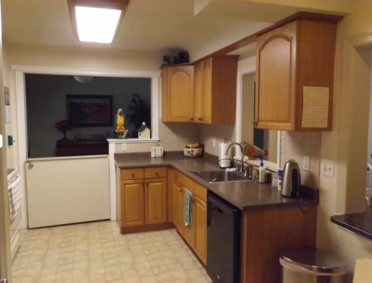 A kitchen with wooden cabinets, a double sink, a dishwasher, and a countertop with various items including a kettle and a paper towel holder. The floor has a light-colored tile pattern, and there is a half-door leading to another room with a decorative parrot figurine visible through the opening.