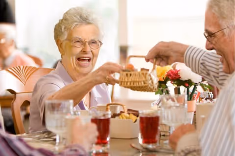 Elderly woman laughing as she reaches for a small basket across a dining table while other seniors enjoy a meal.
