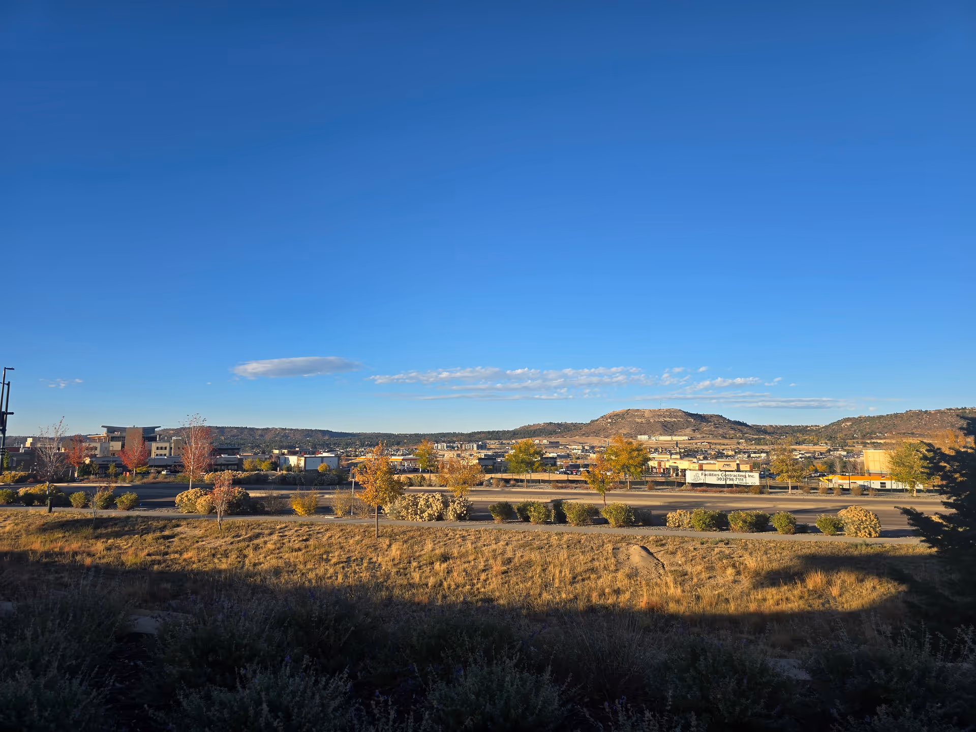 Wide view of a suburban landscape with a road, shrubs, low buildings and distant hills under a clear blue sky.